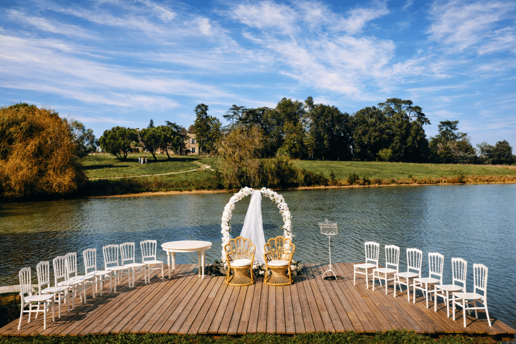 Cérémonie de mariage en extérieur sur une jetée en bois. Deux chaises en rotin et une arche florale face au lac, avec un château en arrière-plan.