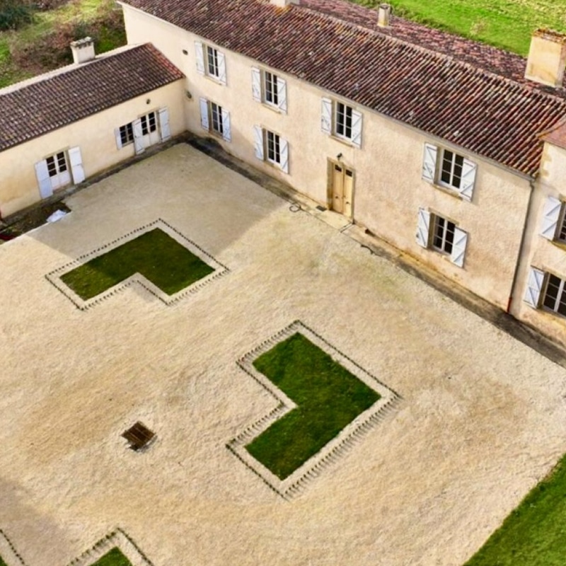 Manoir en pierre beige avec toit en tuiles rouges vu du ciel. Grande cour de gravier aménagée de pelouses vert vif en forme de L.