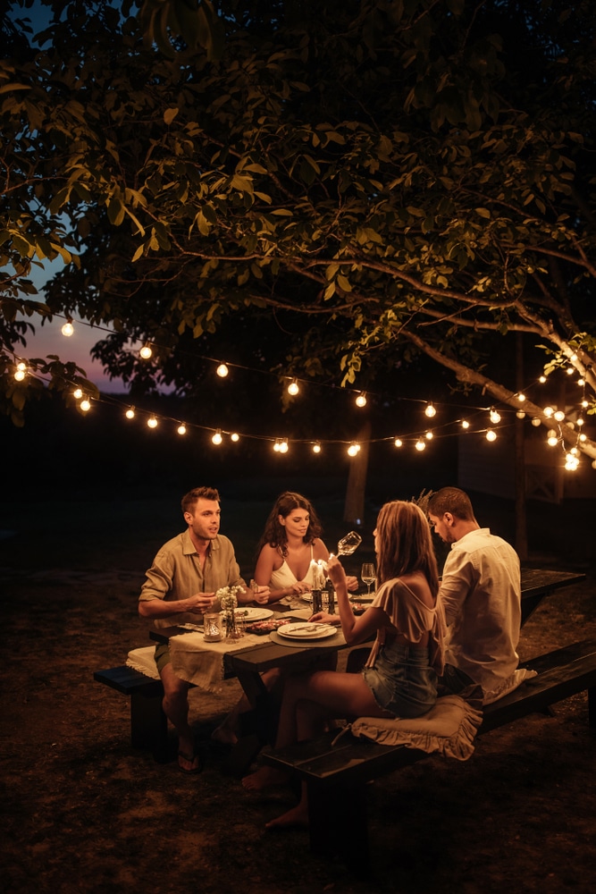 Soirée d'été : Dîner entre amis sous des guirlandes lumineuses. Quatre amis dînent dehors sous un arbre illuminé par des guirlandes lumineuses. Ambiance chaleureuse et conversation joyeuse en soirée d'été.