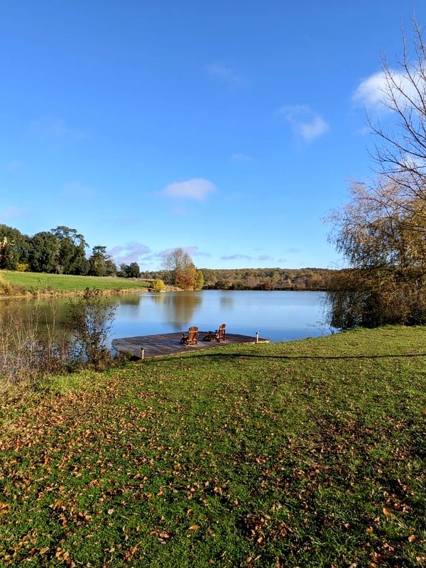 Lac, ponton et chaises : la détente en pleine nature. Ponton en bois et deux chaises Adirondack au bord d'un lac calme, sous un grand ciel bleu d'automne. La rive est couverte d'herbe et de feuilles.