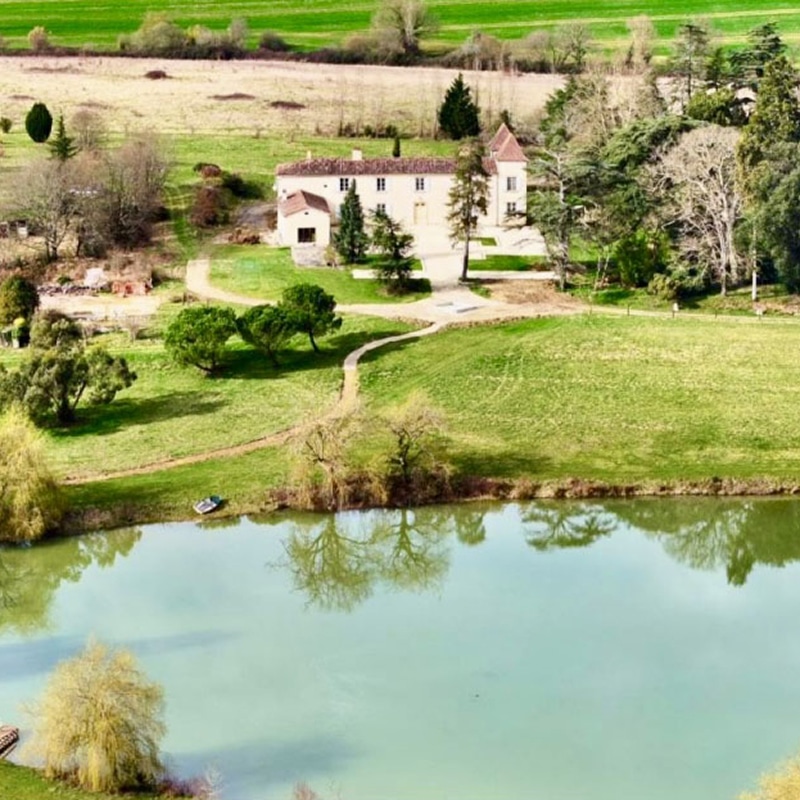 Manoir français en pierre surplombant un étang turquoise et des champs ondulants. Chemins de terre et arbres majestueux.