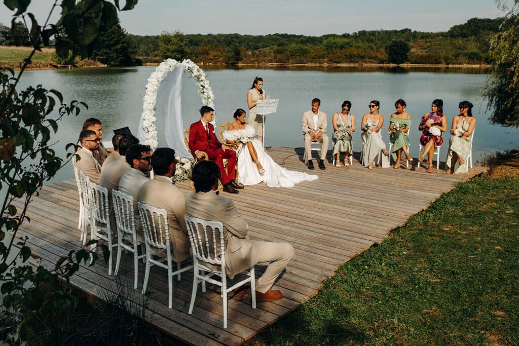 Mariage extérieur chic : cérémonie sur ponton au bord du lac. Cérémonie de mariage extérieur. Couple sur un ponton en bois au bord d'un lac sous une arche florale. Marié en rouge, invités en beige.