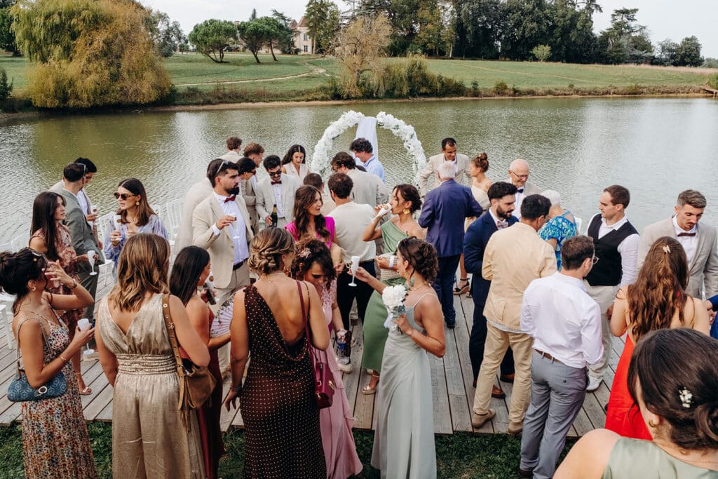 Mariage : cocktail estival élégant sur le ponton du lac. Invités de mariage sur un ponton en bois au bord d'un étang, célébrant lors du cocktail devant une arche florale blanche.