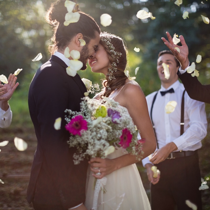 Mariage romantique : couple s'embrassant sous une pluie de pétales Cérémonie de mariage en forêt : les jeunes mariés s'embrassent tendrement sous une pluie de pétales blancs. La mariée tient un bouquet coloré.