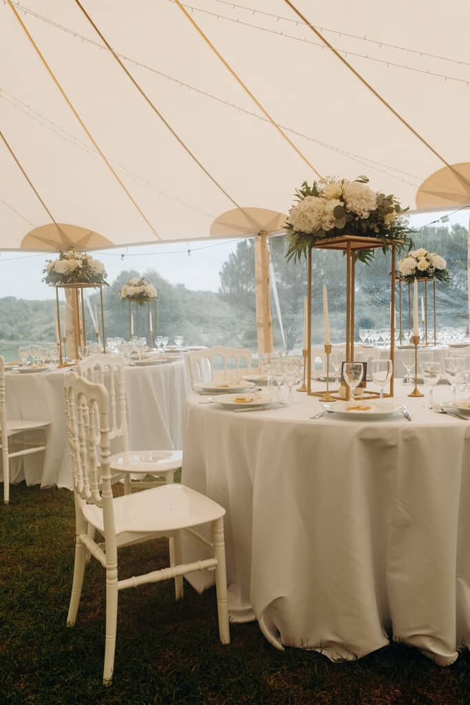 Décoration de mariage : Tables élégantes sous tente champêtre. Tables rondes de réception de mariage sous tente. Décoration élégante: chaises napoléon blanches, centres floraux blancs sur supports dorés.