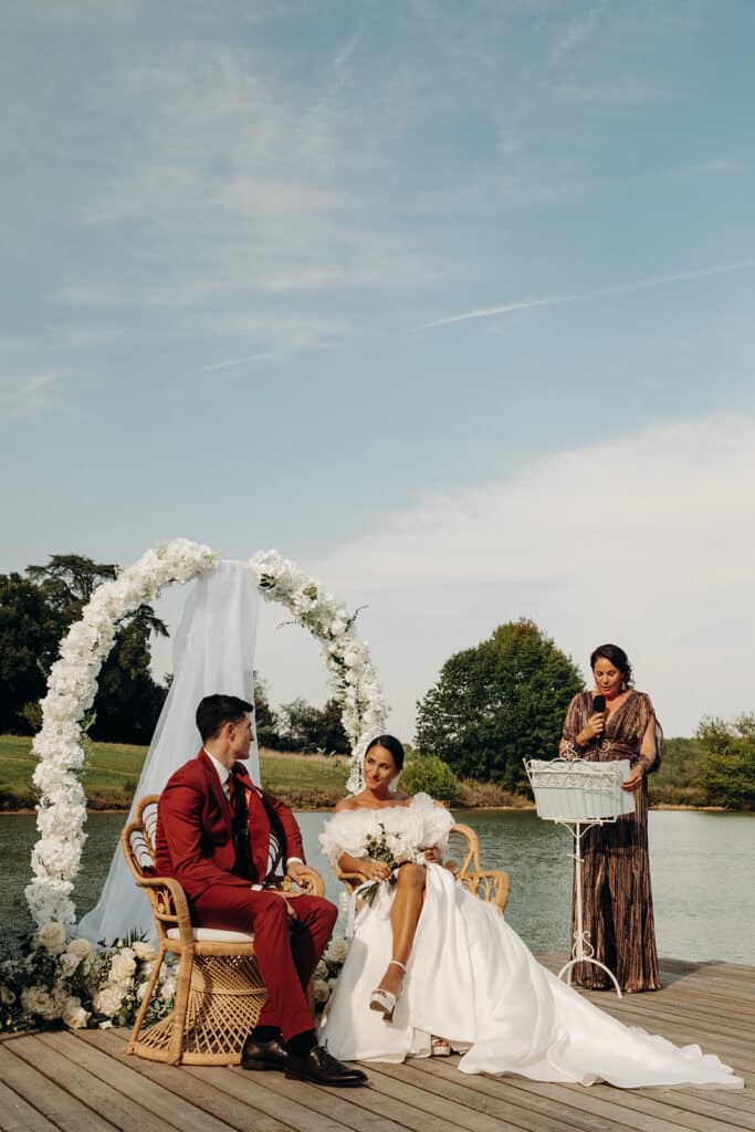 Mariage extérieur : Cérémonie romantique au bord du lac. Mariés assis dans des chaises en osier pendant une cérémonie extérieure. Le marié porte un costume rouge, la mariée une robe blanche bouffante.