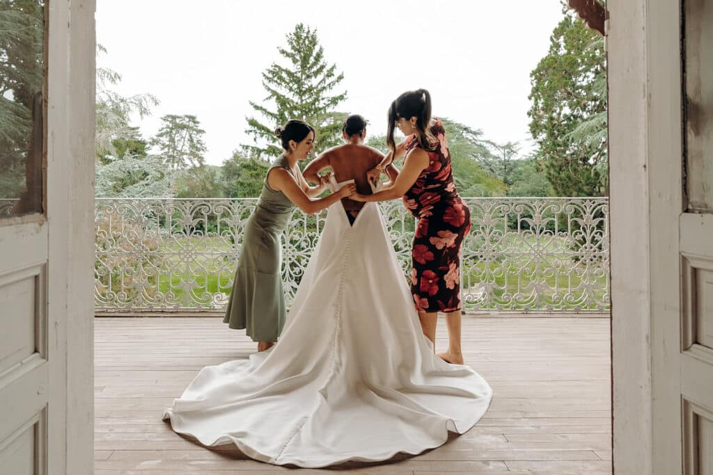 Préparation de la robe de mariée: moment intime sur le balcon. Deux femmes aident la mariée à boutonner sa robe blanche à longue traîne sur un balcon avec balustrade en fer forgé. Vue sur le parc verdoyant.