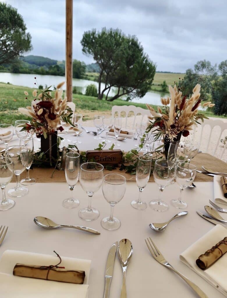 Décoration de Table Mariage Champêtre et Fleurs Séchées Table de mariage champêtre avec fleurs séchées, verres élégants et panneau en bois marqué CHÊNE devant un lac.