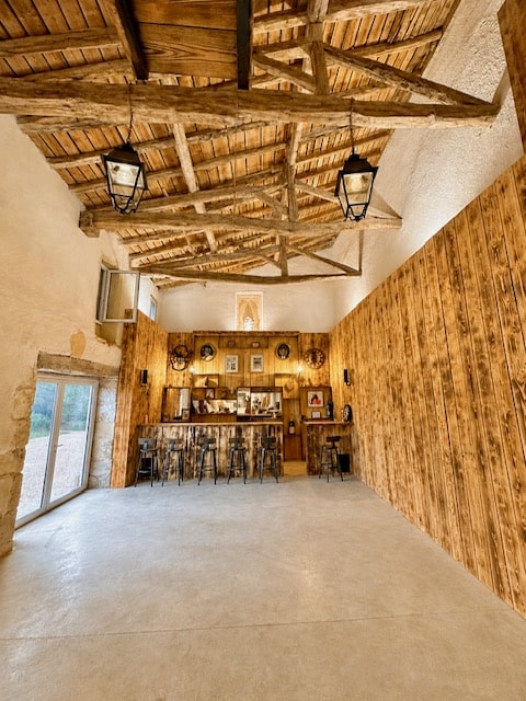 Aménagement Bar Rustique et Charpente Bois de Grange Bar rustique en bois sous charpente apparente dans une grange avec tabourets et luminaires anciens.